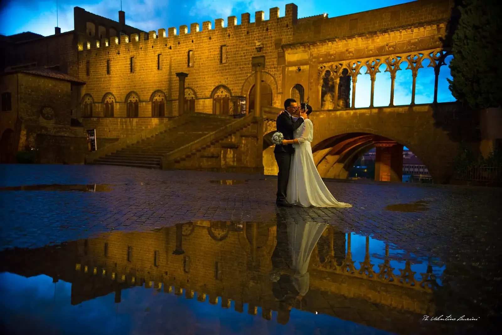 Sposi abbracciati di notte sotto la loggia del Palazzo dei Papi a Viterbo, riflessi nella piazza bagnata